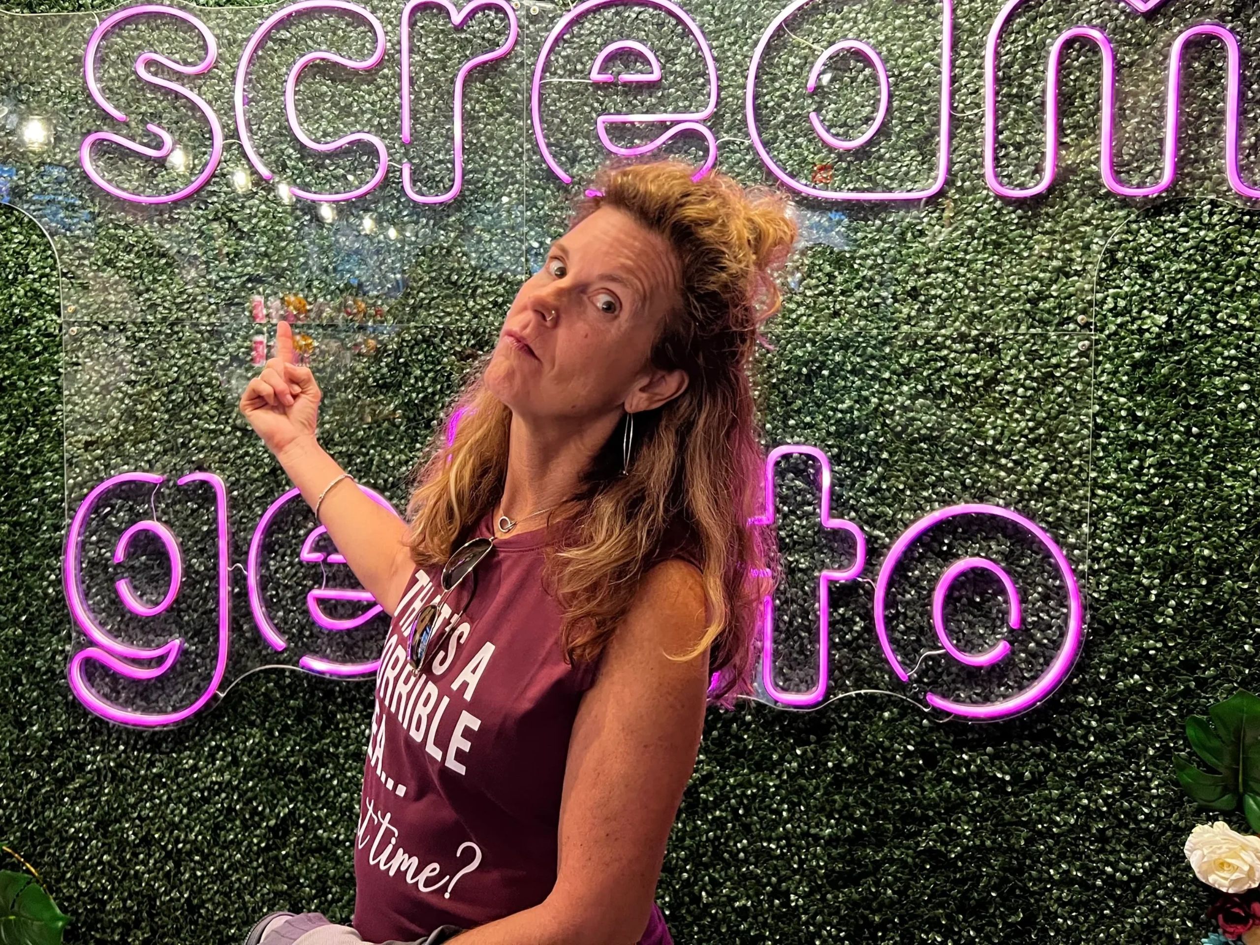Beth Mortell smiling, pointing toward a neon sign with greenery behind her.